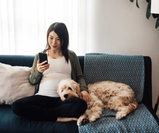 A pregnant woman sits on a couch with her dog while scheduling a virtual doctor's appointment. 
